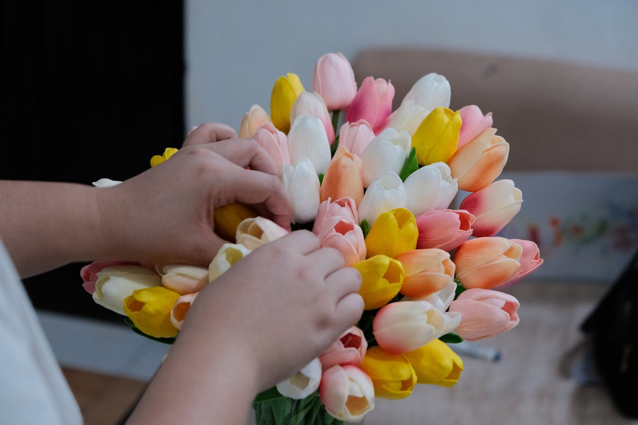 Close-up of hands arranging a colorful bouquet of tulips, including pink, yellow, and white flowers, indoors.